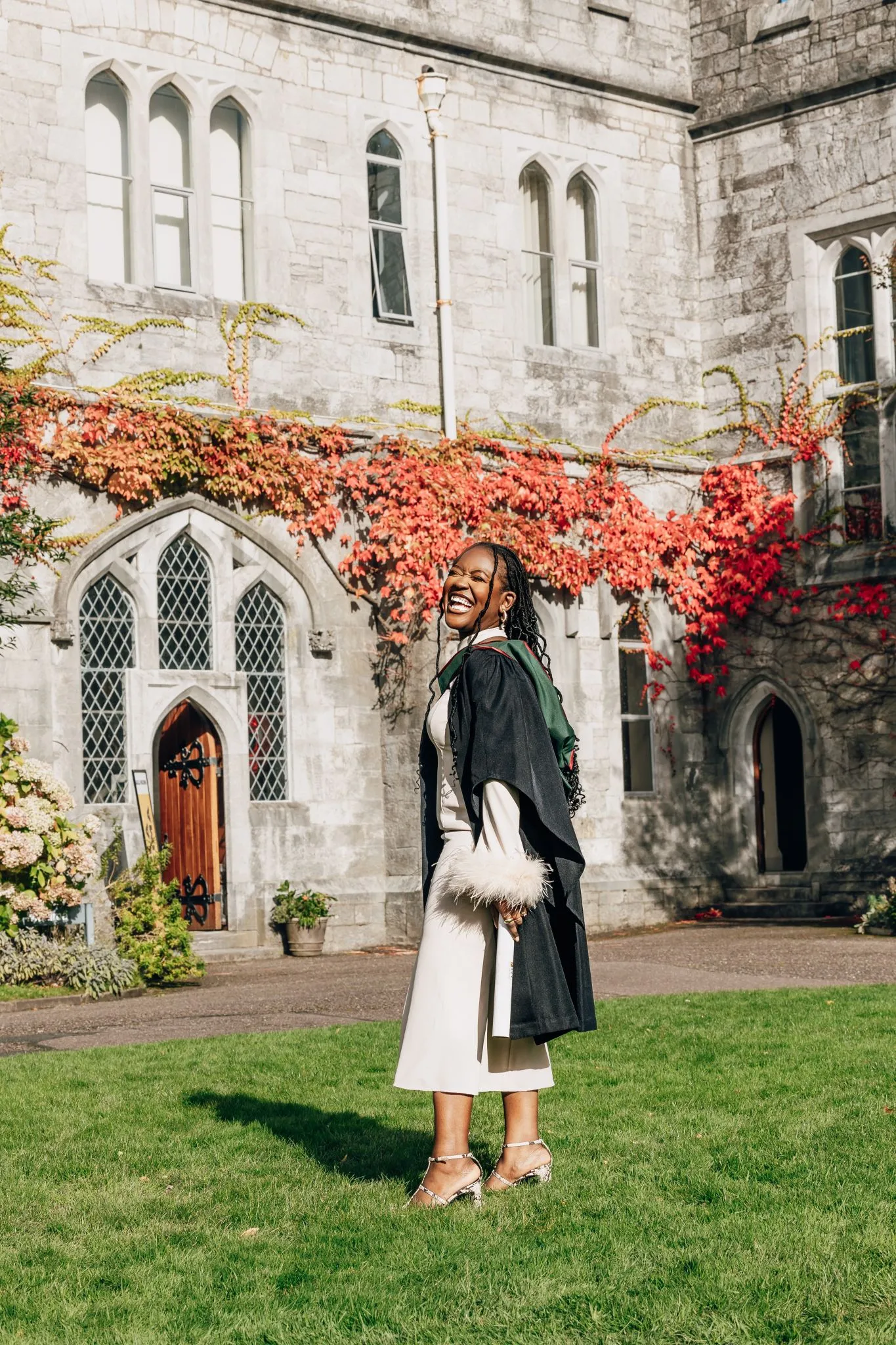 Graduate celebrating in front of historic building with ivy and autumn leaves.