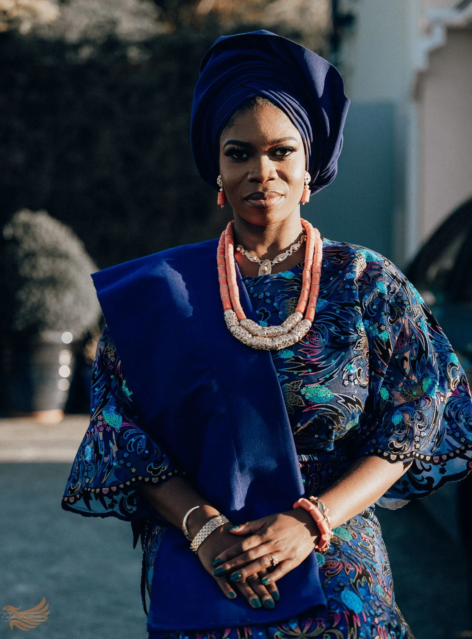 Woman in traditional blue attire with coral beads, standing outdoors.