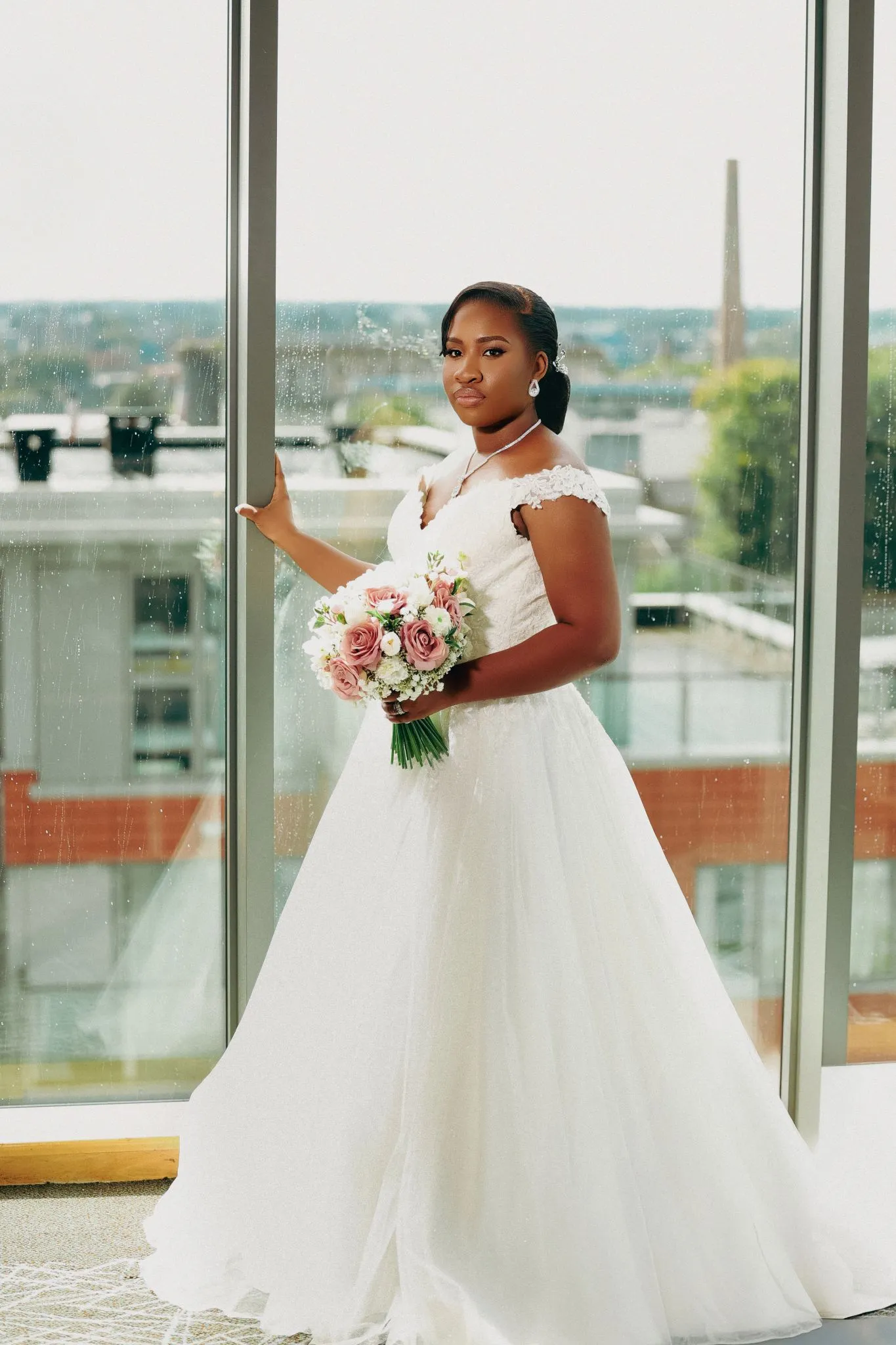 Bride in elegant gown holding a pink and white bouquet by a window.