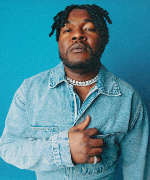 Man wearing a light denim jacket and pearl necklace posing against a blue studio background