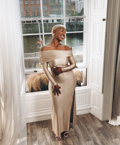 Woman in an off-shoulder beige dress standing by a large window overlooking a canal, smiling in natural light