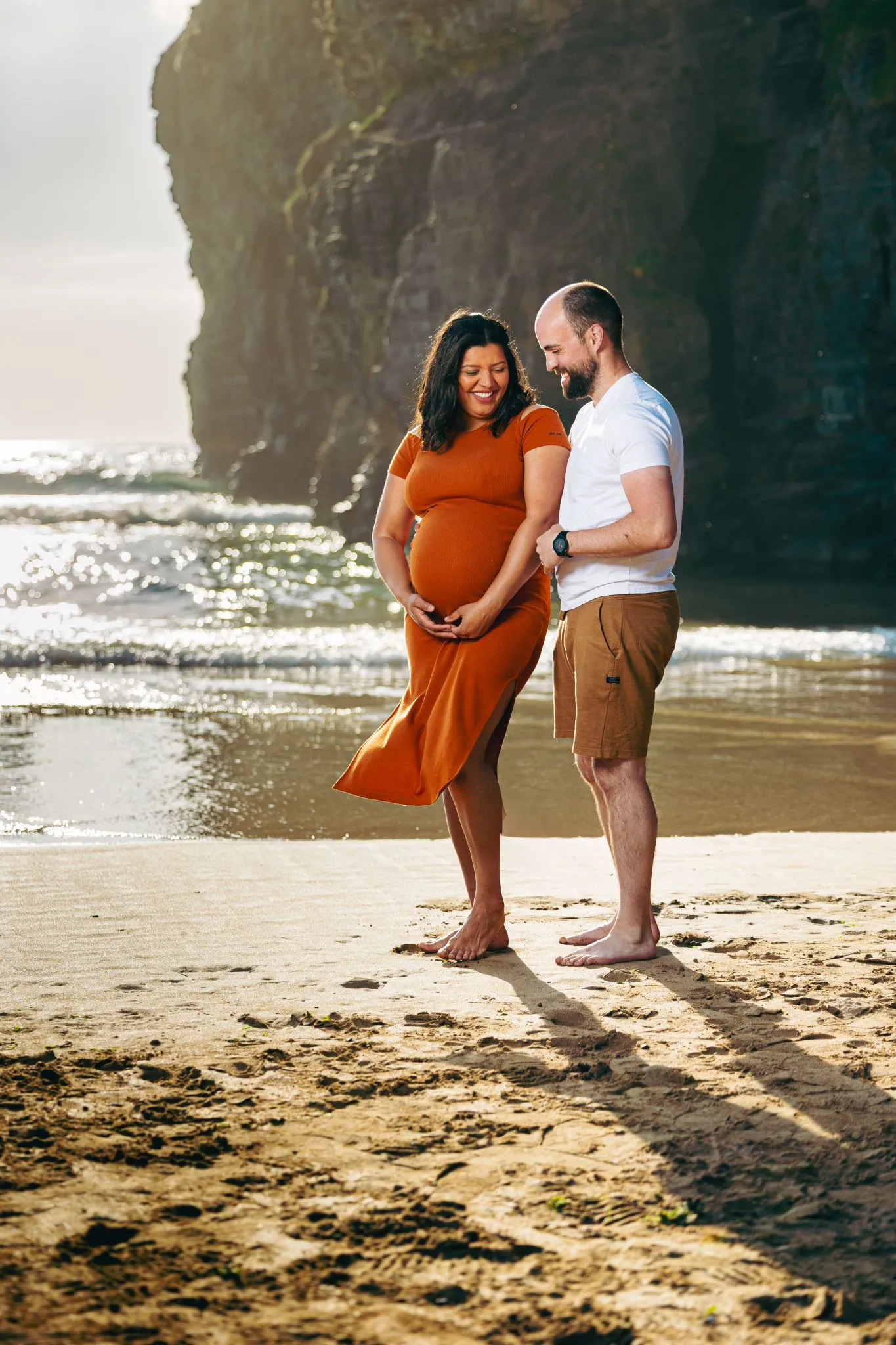 Pregnant couple smiling on a sunny beach with waves and cliffs.