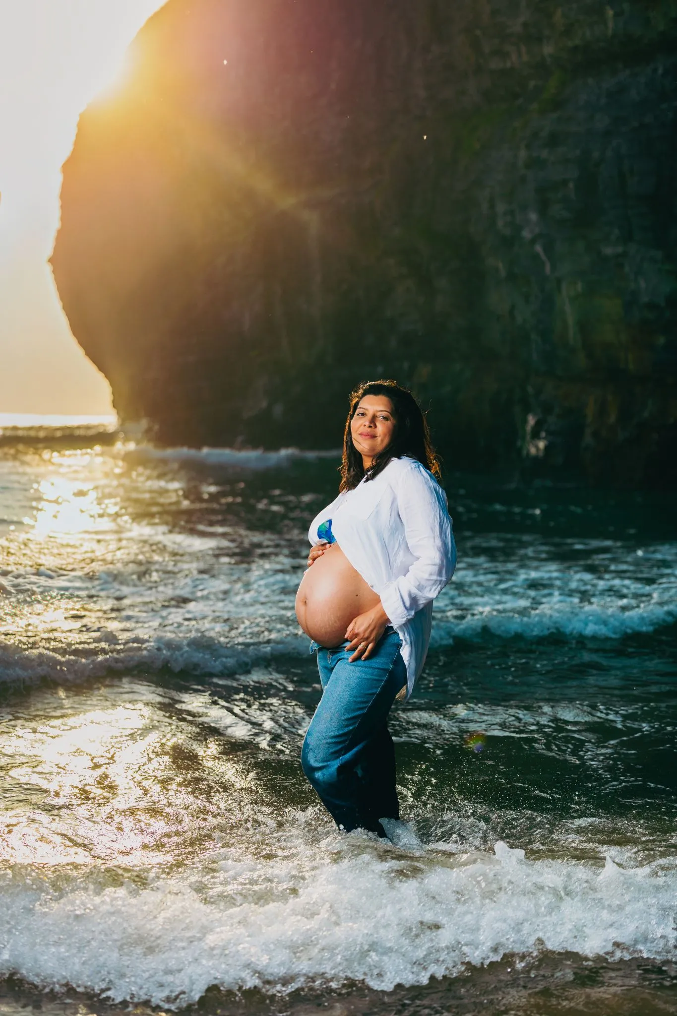 Pregnant woman standing on a beach at sunset with waves around her.