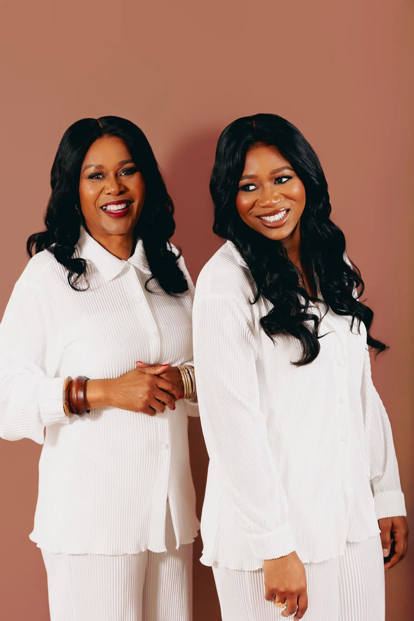 Two smiling women in white outfits against a warm background.