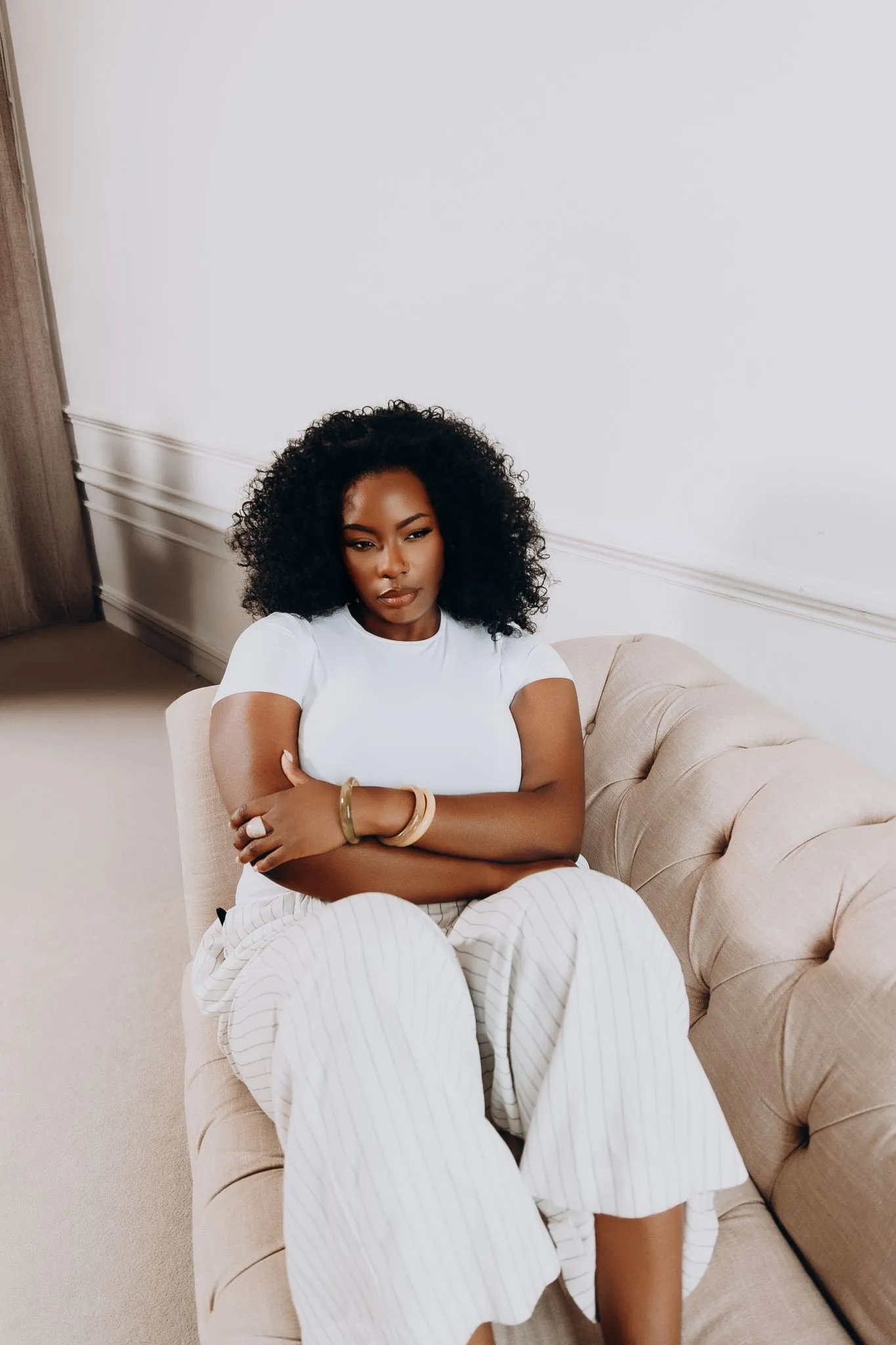 Woman sitting on a beige couch with arms crossed, looking thoughtful.